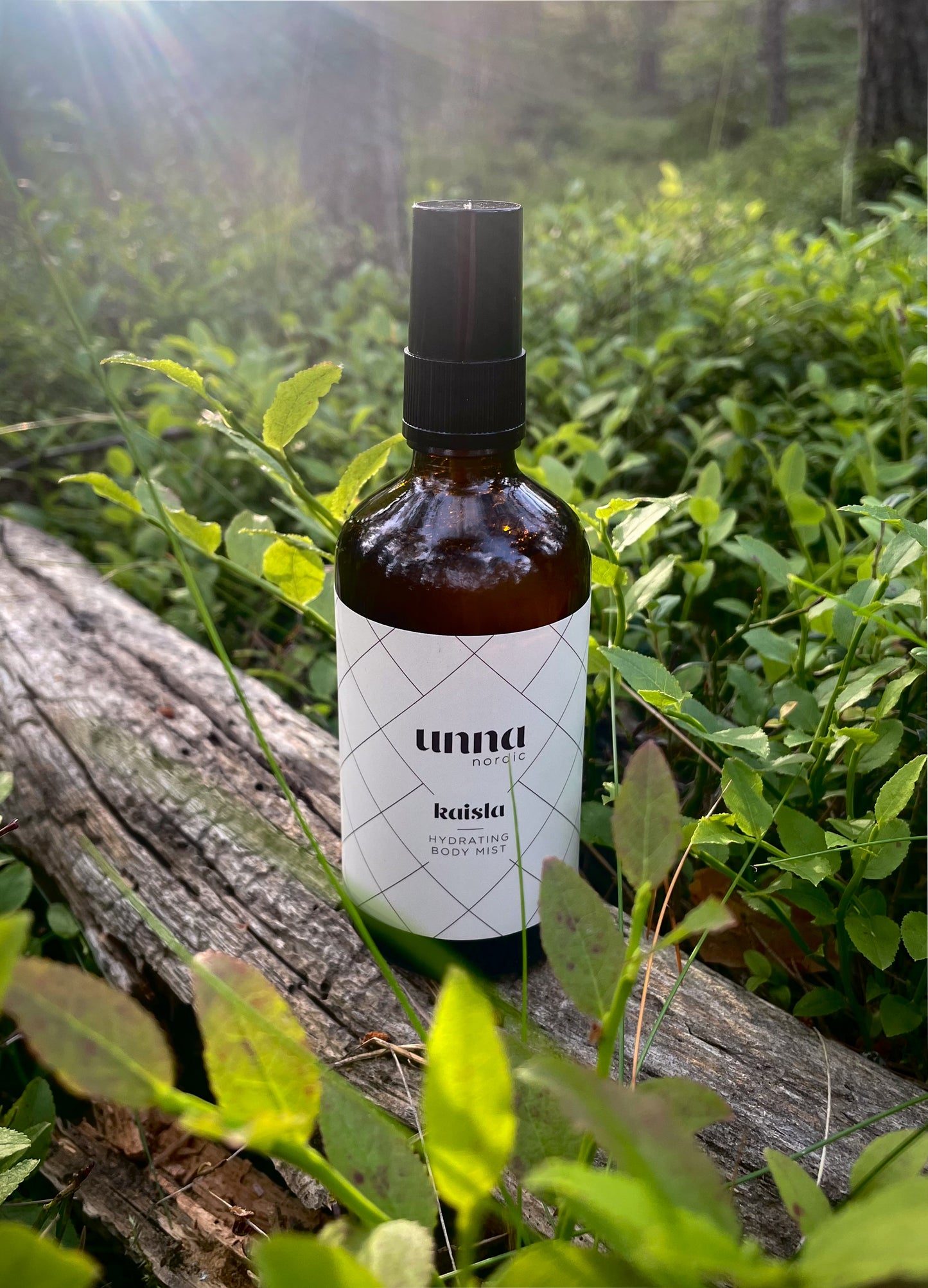 Kaisla Hydrating Body Mist hoitosuihke ruskeassa lasipullossa kuvattuna kesäisessä metsässä puunrungolla ilta-auringossa