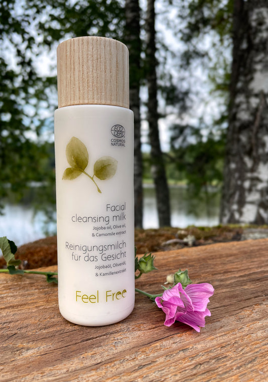 Feel Free Facial Cleansing Milk - Puhdistusmaito 250ml pullo kuvattuna järven rannalla puisen tason päällä vaaleanpunaisen kukan vieressä