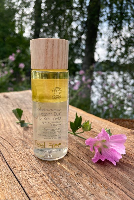 puukorkkinen Feel Freen kaksifaasinen Instant Duo Makeup Remover pullo kuvattuna vanhalla laudalla järven rannalla, vieressä taustalla kasvava vaaleanpunainen kukka.